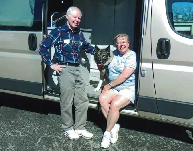 The Whitmans and their dog rest in the doorway of their silver Sportsmobile ProMaster camper conversion.